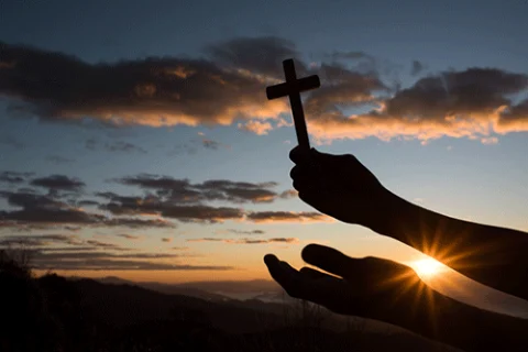mãos segurando uma cruz remetendo a fé no pôr do sol