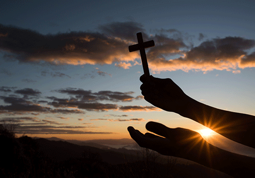 mãos segurando uma cruz remetendo a fé no pôr do sol